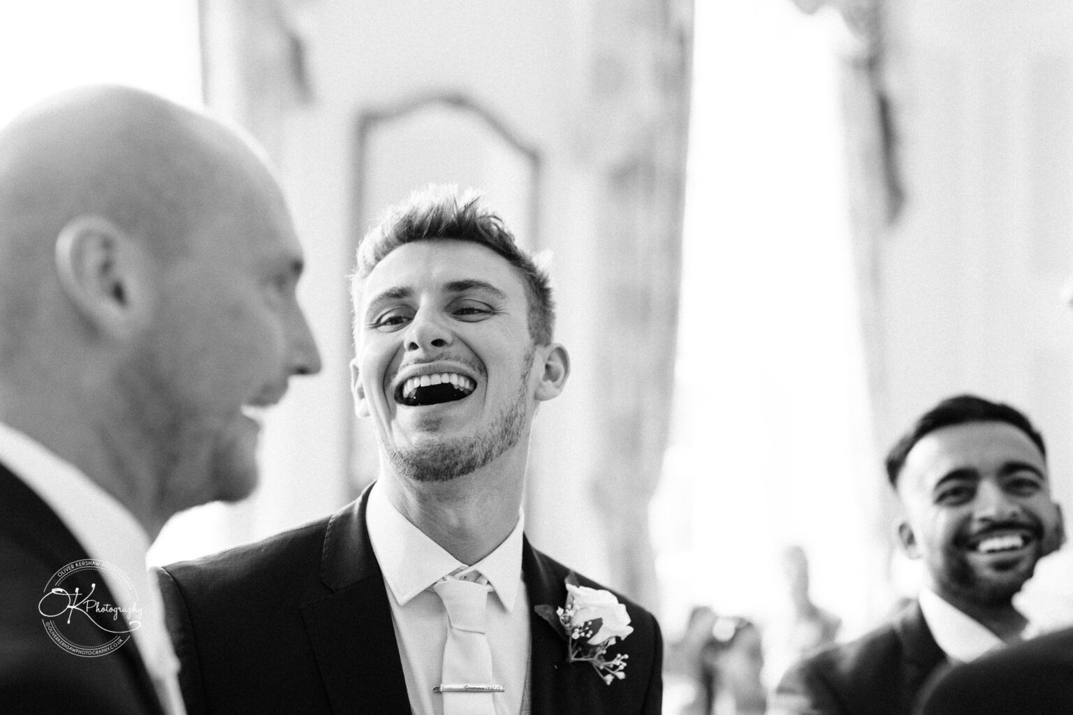 Three men in formal attire laughing and conversing at an indoor event, one man wearing a flower boutonnière.