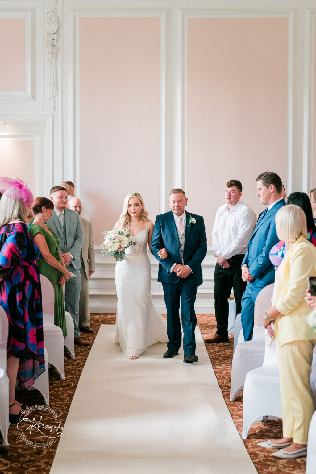 Bride in a white wedding dress walking down the aisle with her father, surrounded by guests seated in an elegant room with light pink and white decor.