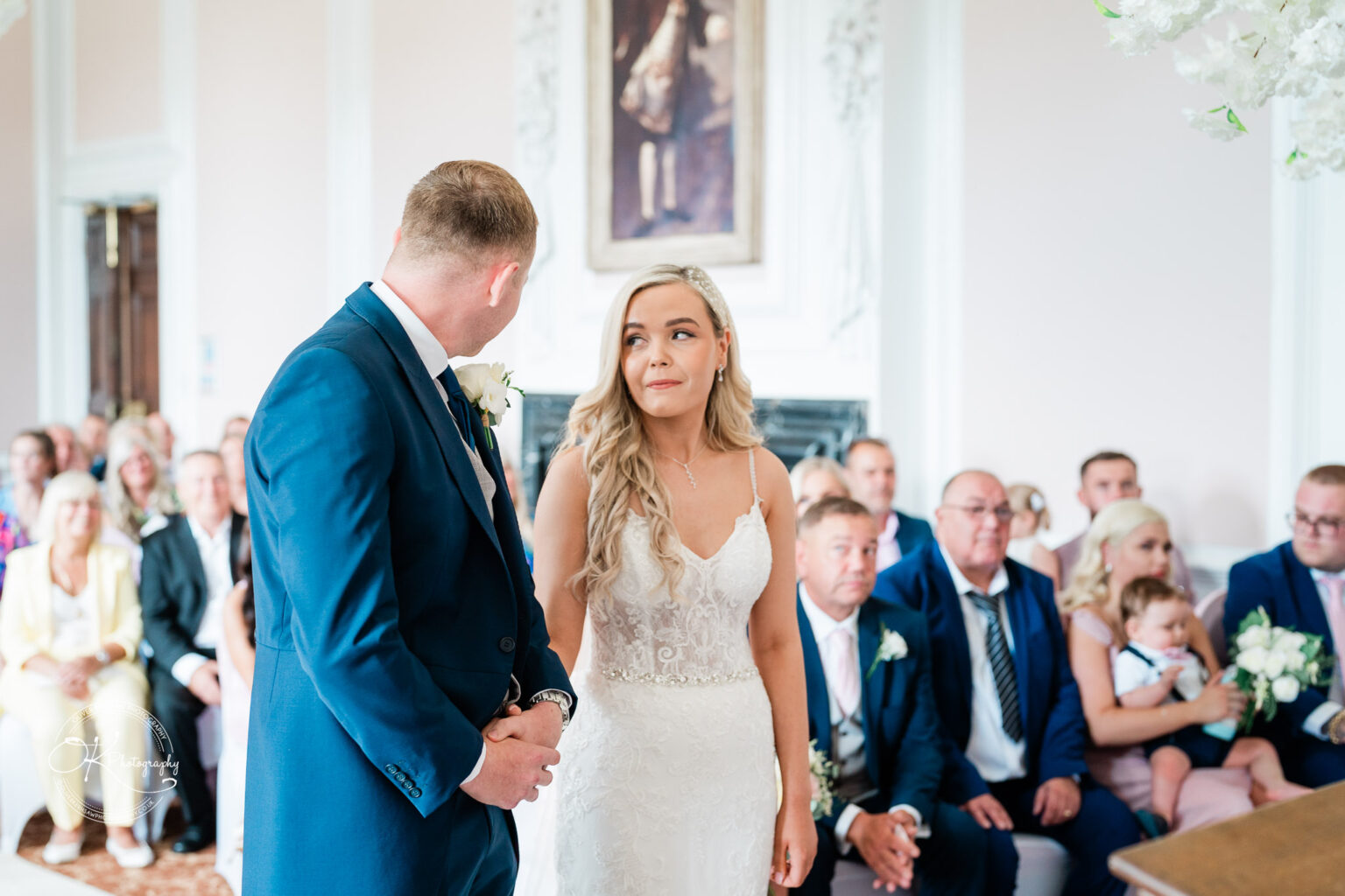A couple stands facing each other during a wedding ceremony in a bright room with guests seated in the background.