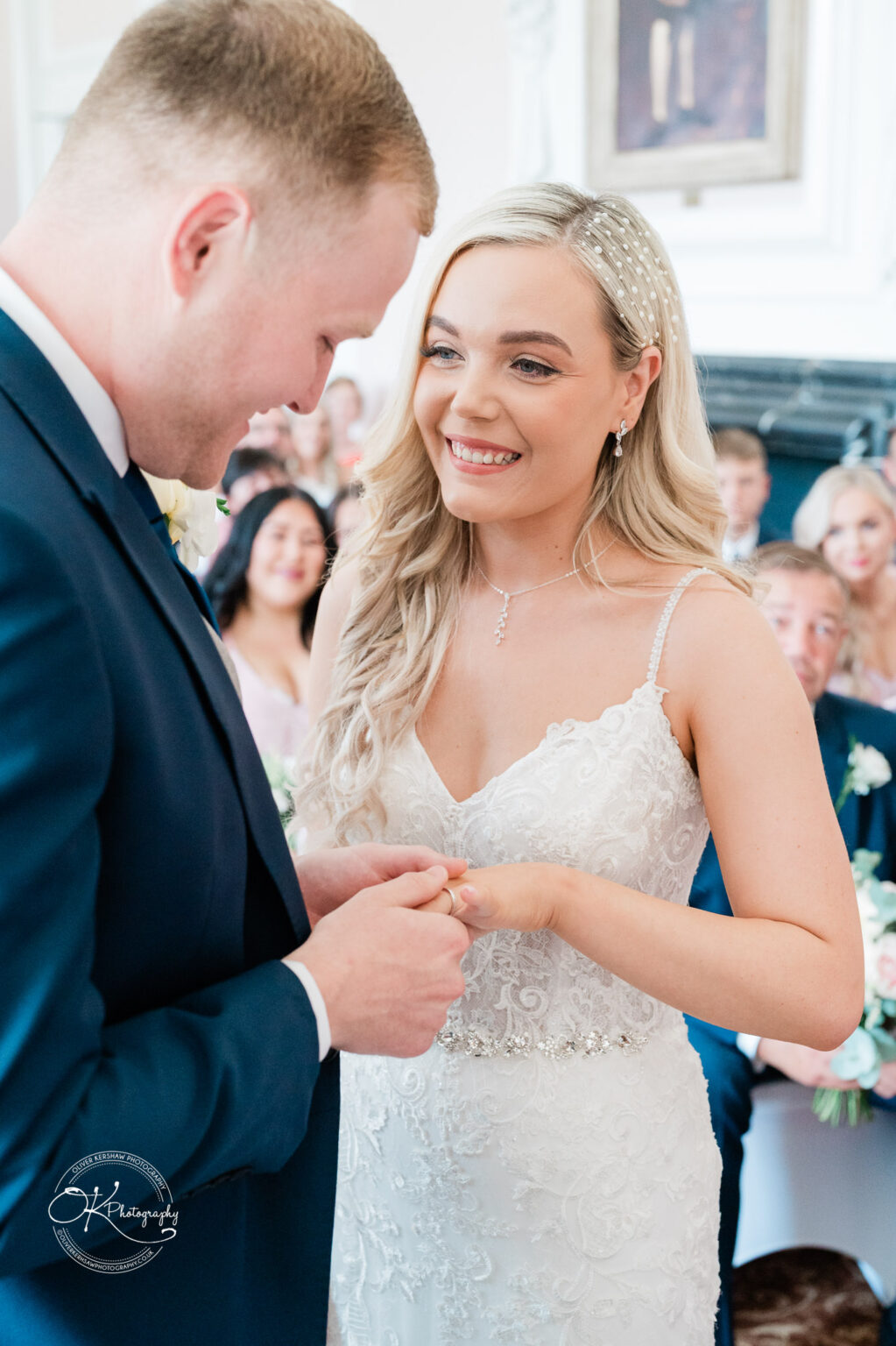 A bride and groom exchanging rings during a wedding ceremony, with guests seated in the background.