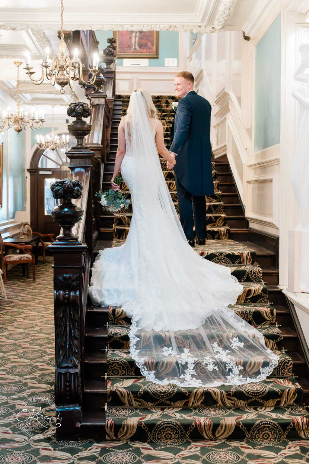 A bride and groom hold hands while walking up a grand staircase at Bosworth Hall Hotel, with ornate chandeliers and elegant decor in the background.