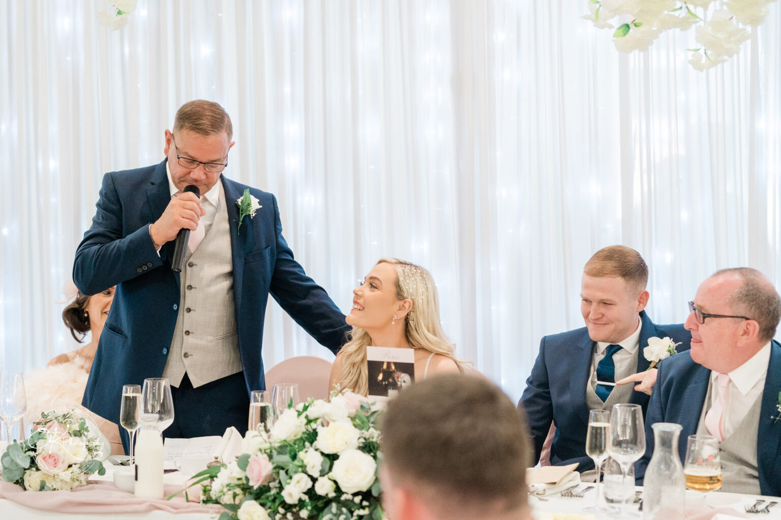 A wedding speech being given by a man in a suit to a seated bride with guests at Bosworth Hall Hotel, with floral decorations and a white backdrop.