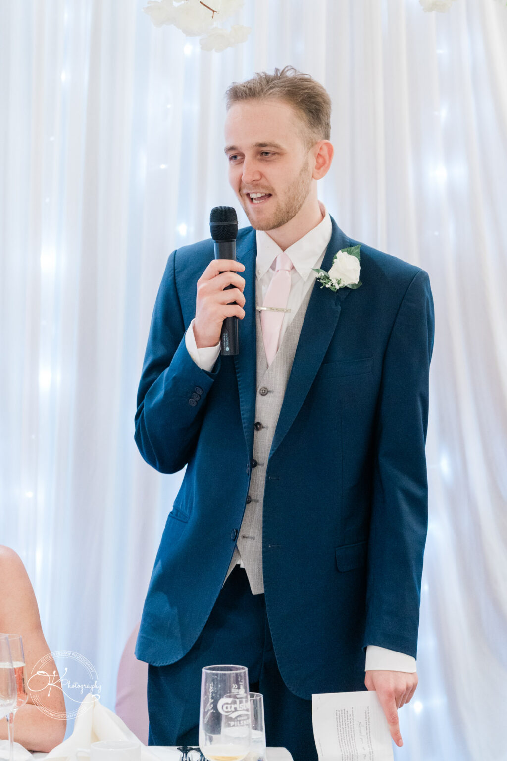 A man in a blue suit and pink tie holding a microphone, giving a speech.