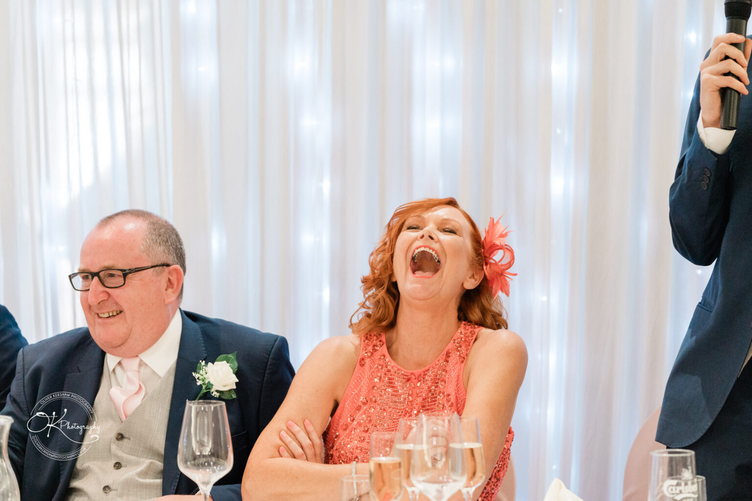Man and woman seated and laughing at a formal event, with a person to the right holding a microphone, against a backdrop of white drapes with fairy lights.