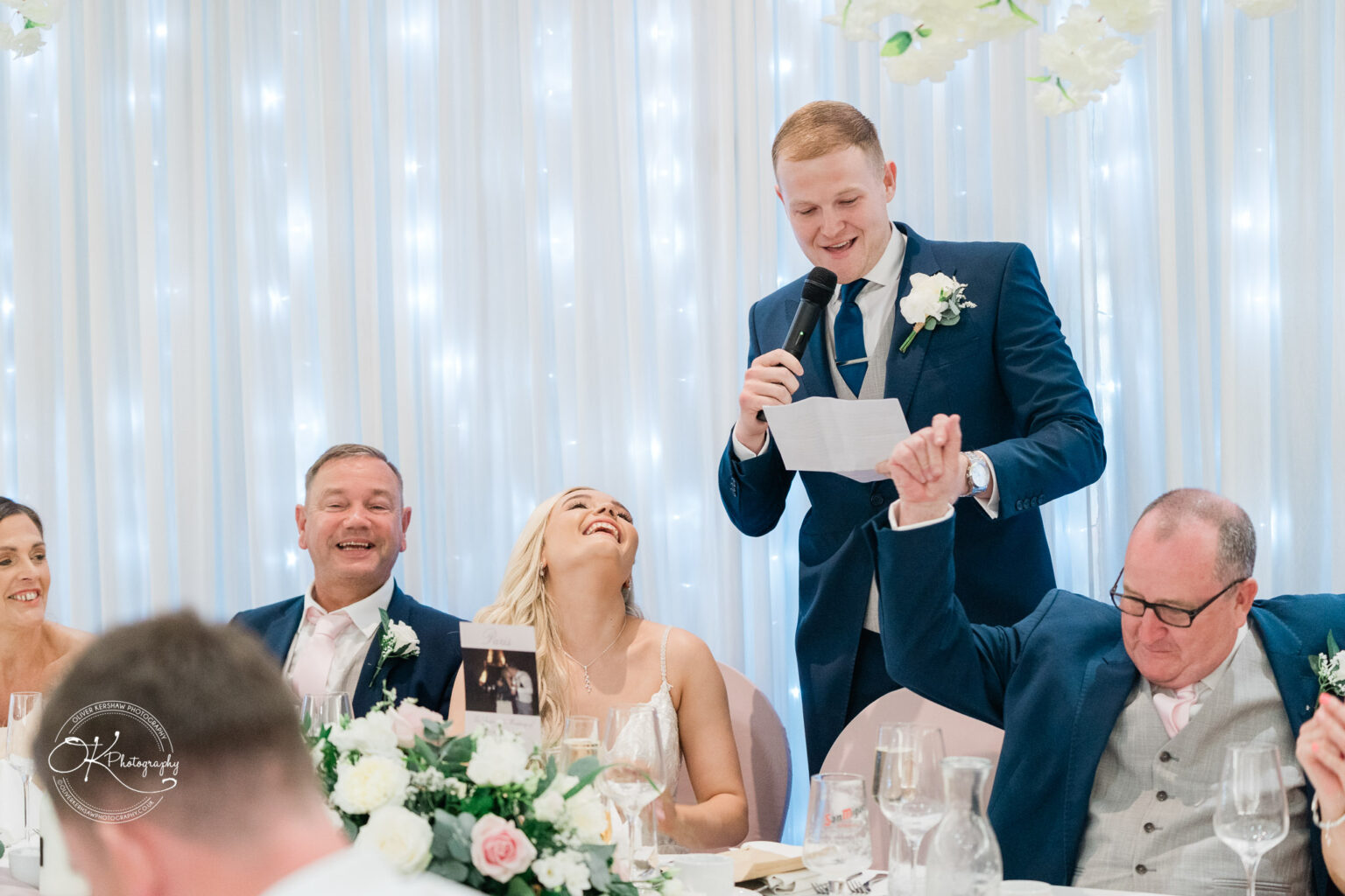 A wedding speech being delivered by a man in a blue suit at a reception, with a bride laughing, a groom smiling, and two other guests sitting at a table with floral decorations.