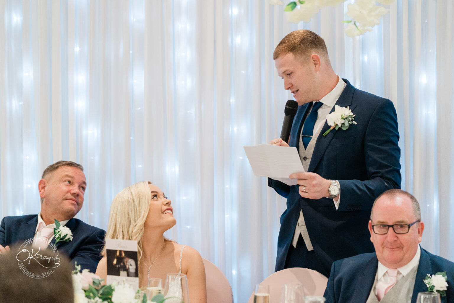 Wedding reception with a man in a suit giving a speech while others sit and listen.