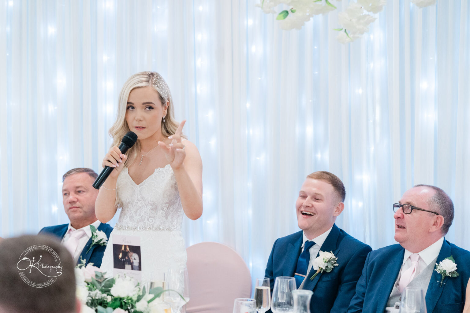 A bride gives a speech at her wedding reception while three groomsmen seated next to her smile.