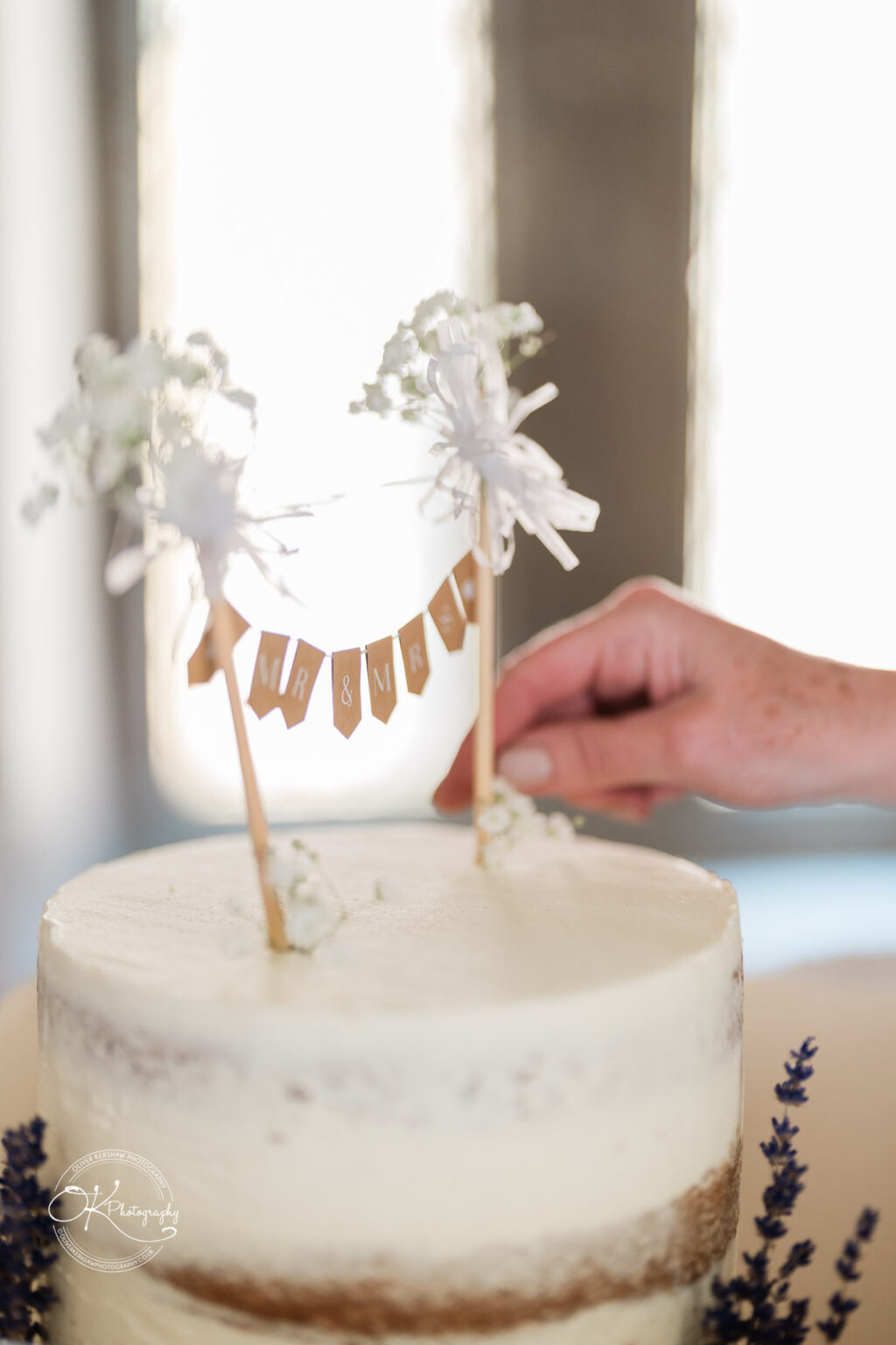 Rothley Court Hotel wedding photography A hand adjusting a "Mr & Mrs" cake topper on a white frosted, semi-naked cake decorated with small flowers.