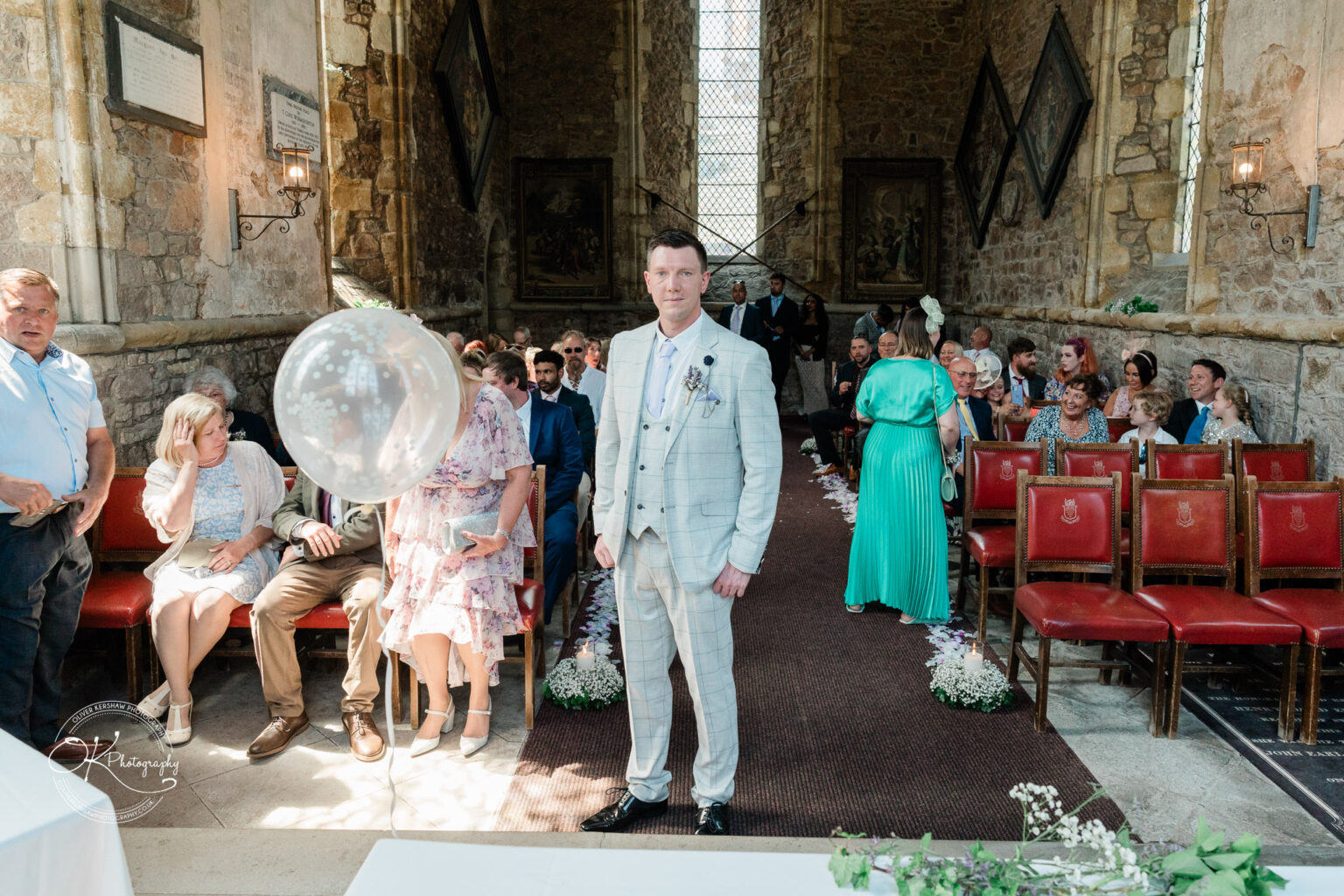 Rothley Court Hotel wedding photography Man in a grey suit standing in front of seated guests in a stone-walled church.