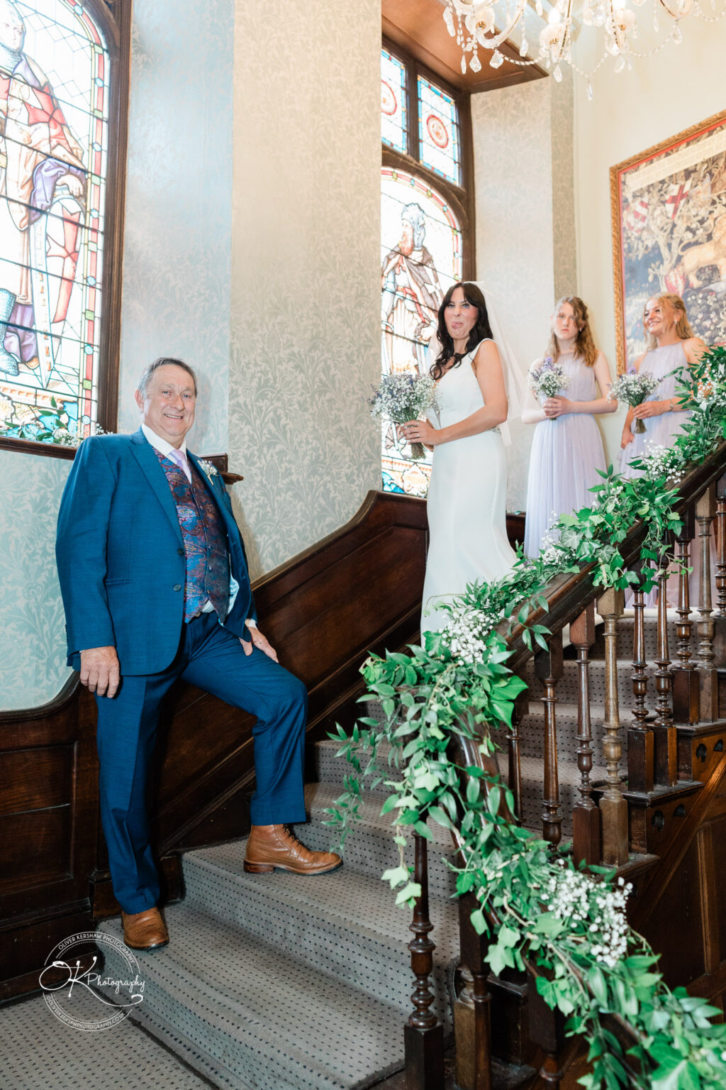 Rothley Court Hotel wedding photography A man in a blue suit stands at the foot of a staircase decorated with greenery; a bride in a white dress and three bridesmaids in light dresses stand on the stairs with stained glass windows in the background.