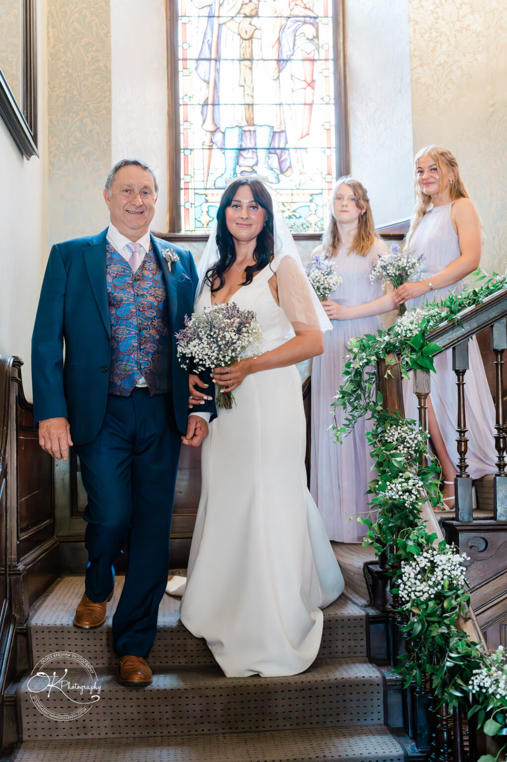 Rothley Court Hotel wedding photography A bride in a white gown and veil walks down the stairs holding hands with an older man in a suit, accompanied by three bridesmaids in lilac dresses, holding bouquets. The stairway is decorated with green and white flowers, and a stained glass window is visible in the background.