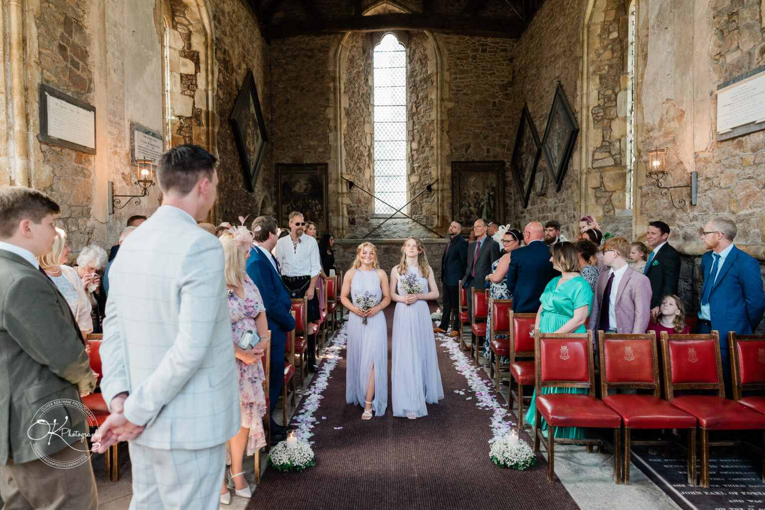 Rothley Court Hotel wedding photography Two bridesmaids in lavender dresses walking down the aisle of a stone-walled church, surrounded by seated guests.