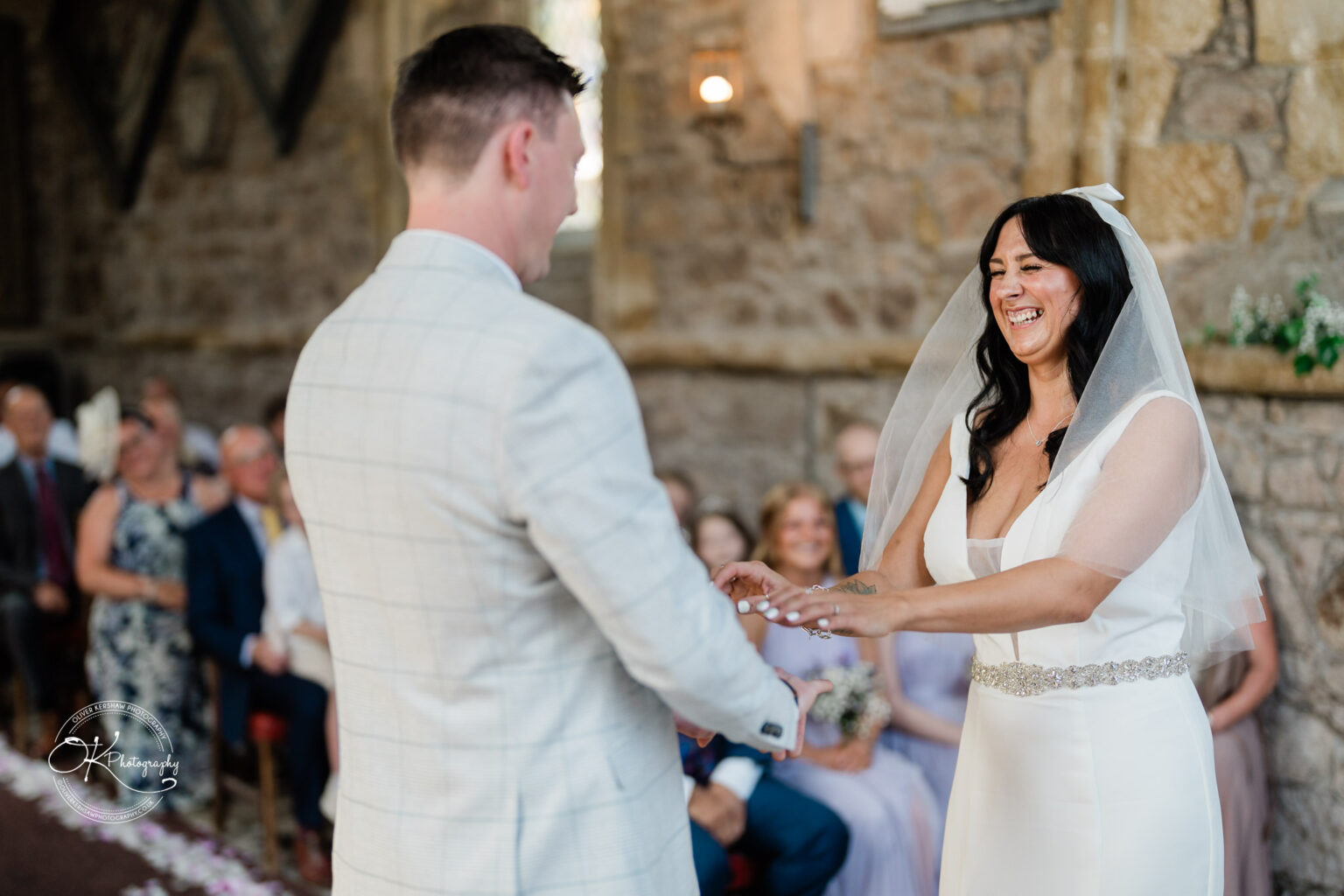 Rothley Court Hotel wedding photography A bride and groom smiling at each other during their wedding ceremony, with seated guests in the background.