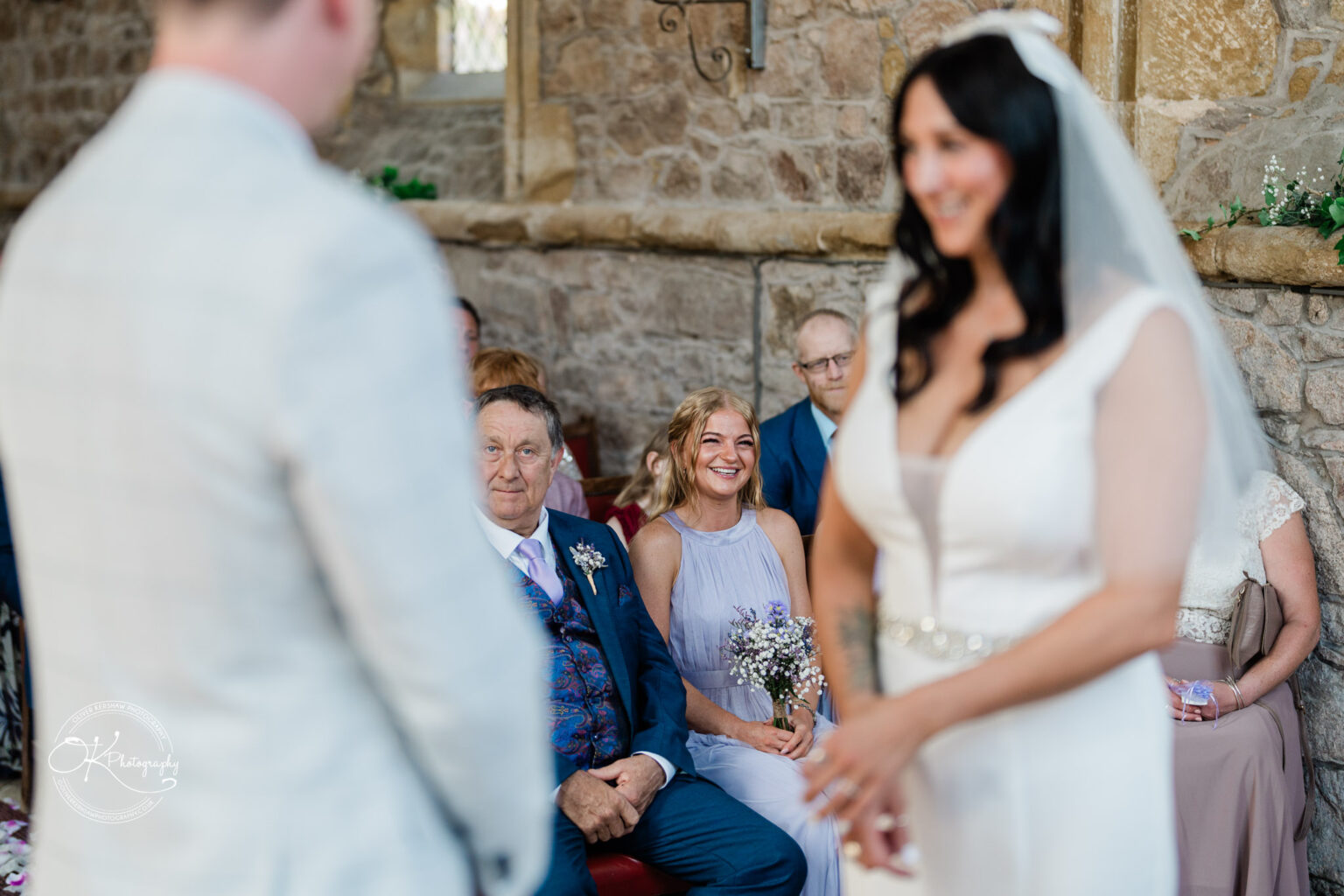 Rothley Court Hotel wedding photography Bride and groom during their wedding ceremony, with guests including a smiling bridesmaid and others seated inside a stone-walled venue.