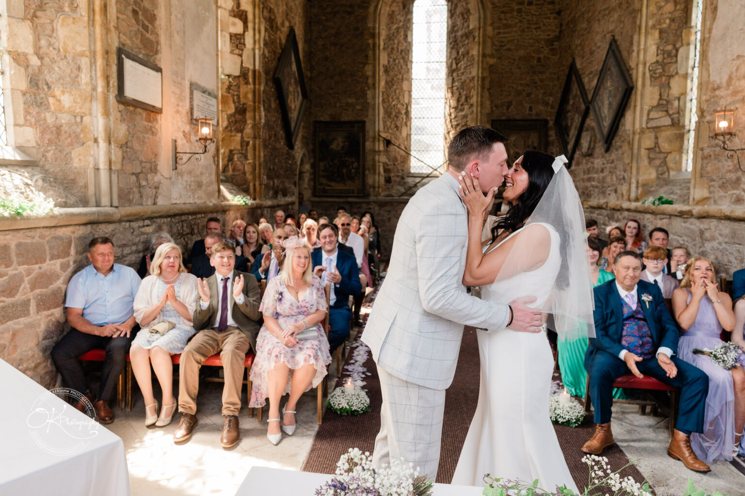 Rothley Court Hotel wedding photography Bride and groom kissing in a historic stone church, surrounded by smiling and applauding guests.