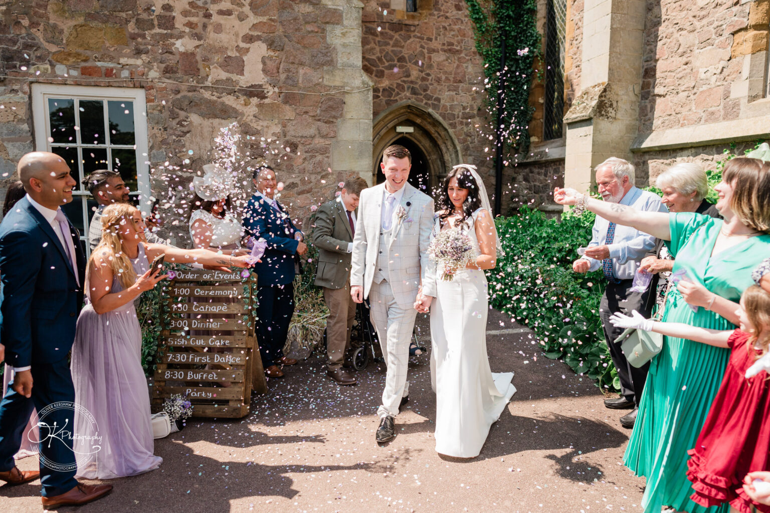 Rothley Court Hotel wedding photography Bride and groom walking outside a stone building, surrounded by guests throwing confetti, with an order of events board in the background.