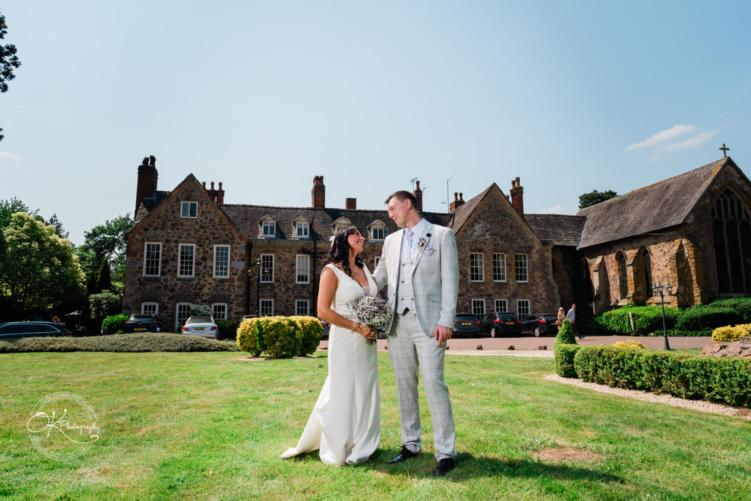 Rothley Court Hotel wedding photography A newlywed couple standing on a lawn, with a historic stone building and a small chapel in the background.