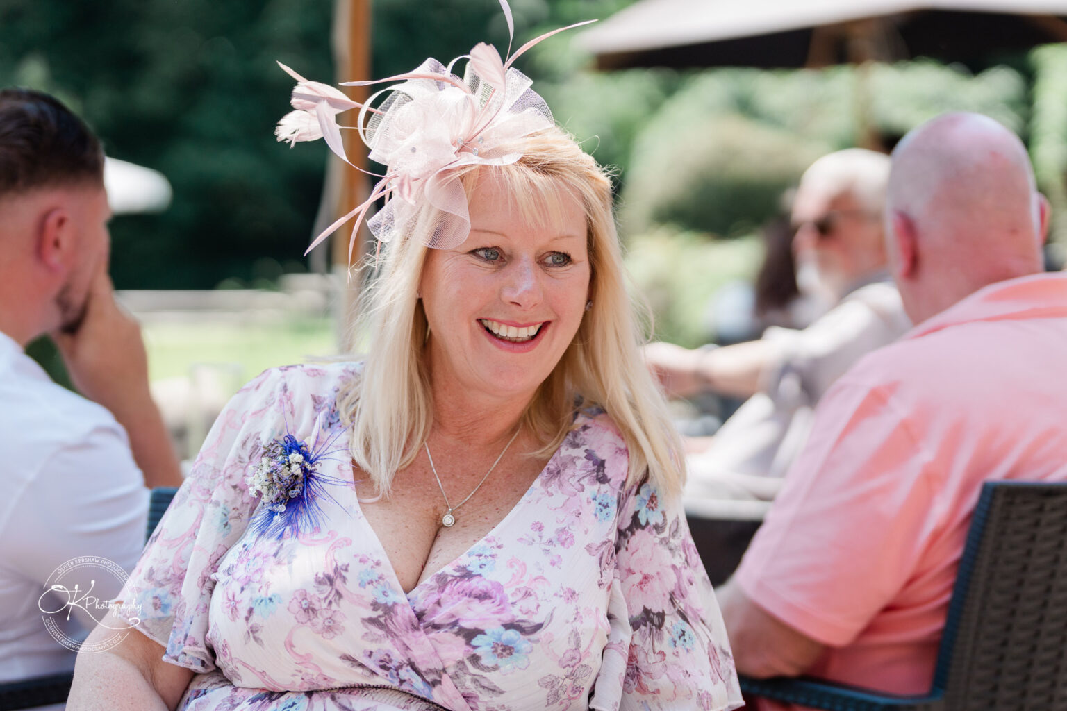 Rothley Court Hotel wedding photography Woman in a floral dress with a pink fascinator, smiling at an outdoor event.