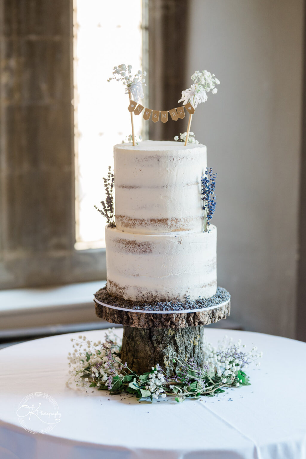 Two-tiered semi-naked wedding cake with "Mr & Mrs" bunting topper, decorated with sprigs of baby’s breath and lavender, displayed on a rustic wooden stand.