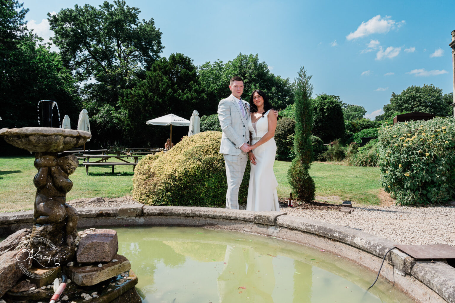 Rothley Court Hotel wedding photography A couple standing by a small fountain in a lush garden under a clear blue sky.