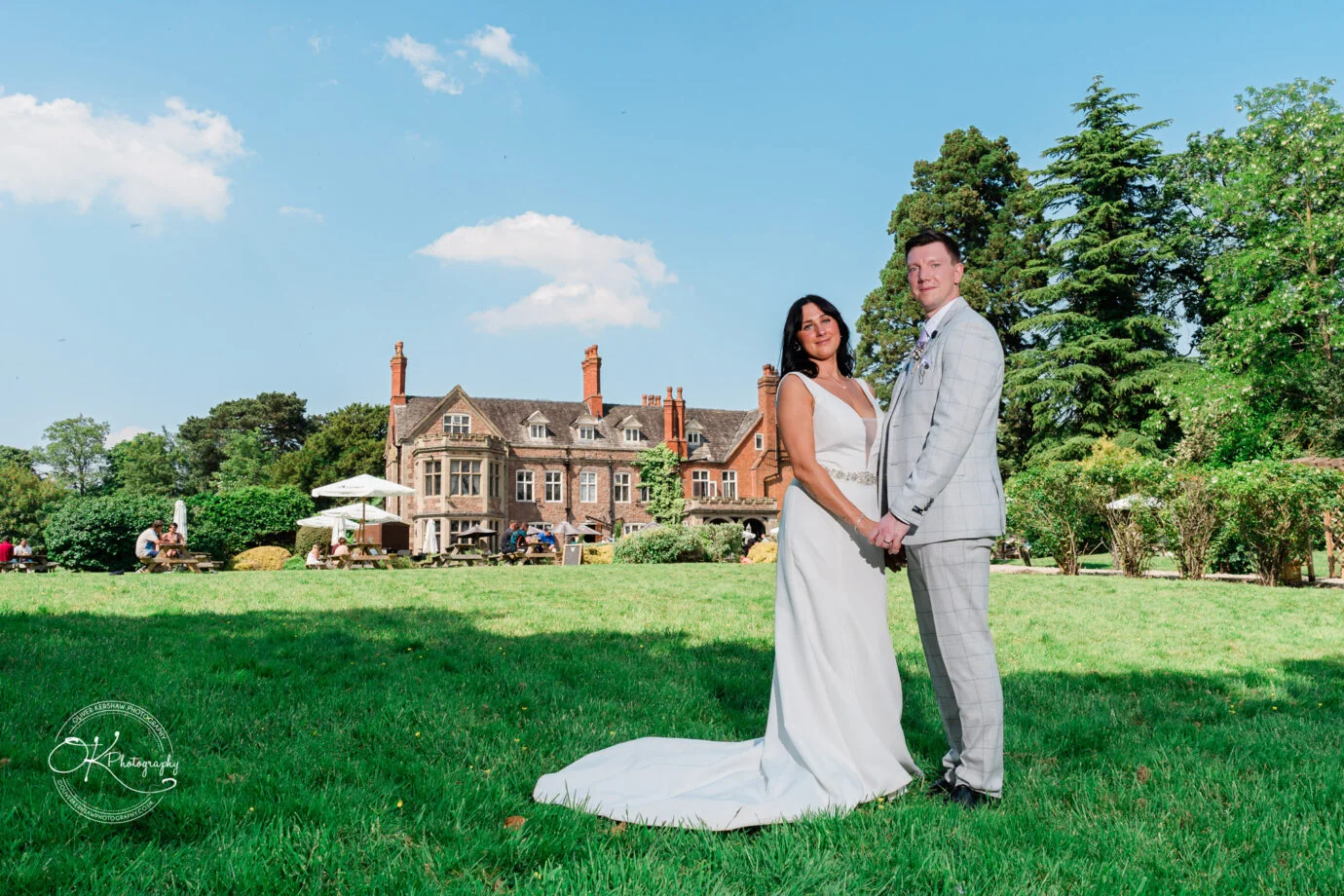 Wedding couple in a garden standing in front of a grand historic building on a sunny day.