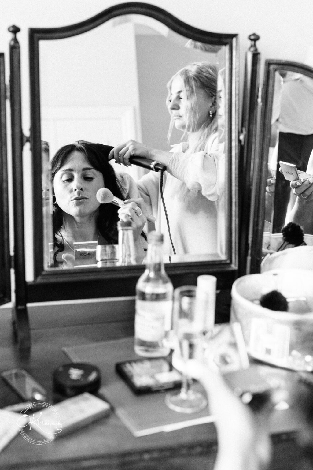Rothley Court Hotel wedding photography Black and white photo of a woman seen through a mirror being styled and applying makeup, with various cosmetic items and a drink on the table.