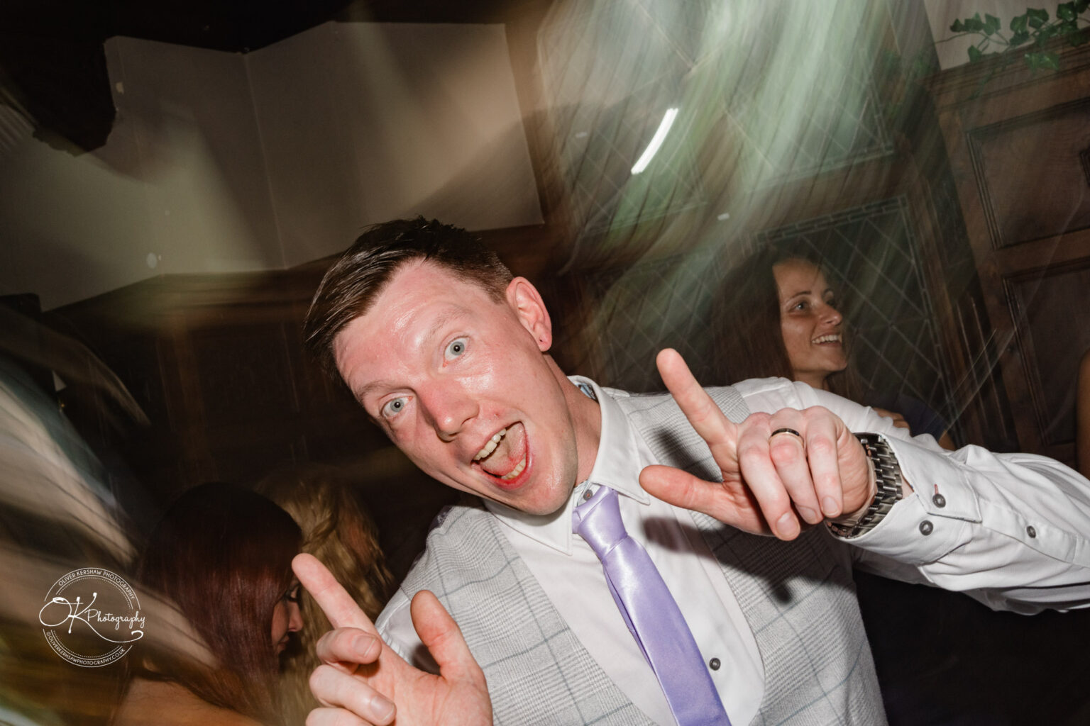 Rothley Court Hotel wedding photography Man in a white shirt and grey vest with a purple tie making a playful expression and pointing upwards with both hands in a blurred background party setting.