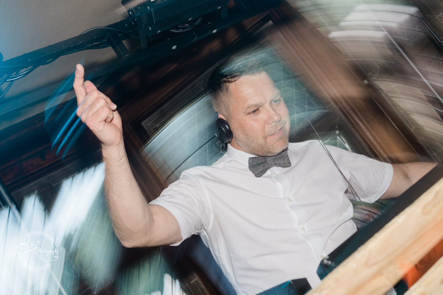A DJ wearing headphones and a bow tie, raising one hand while working at a DJ booth, with streaks of light creating a dynamic effect.