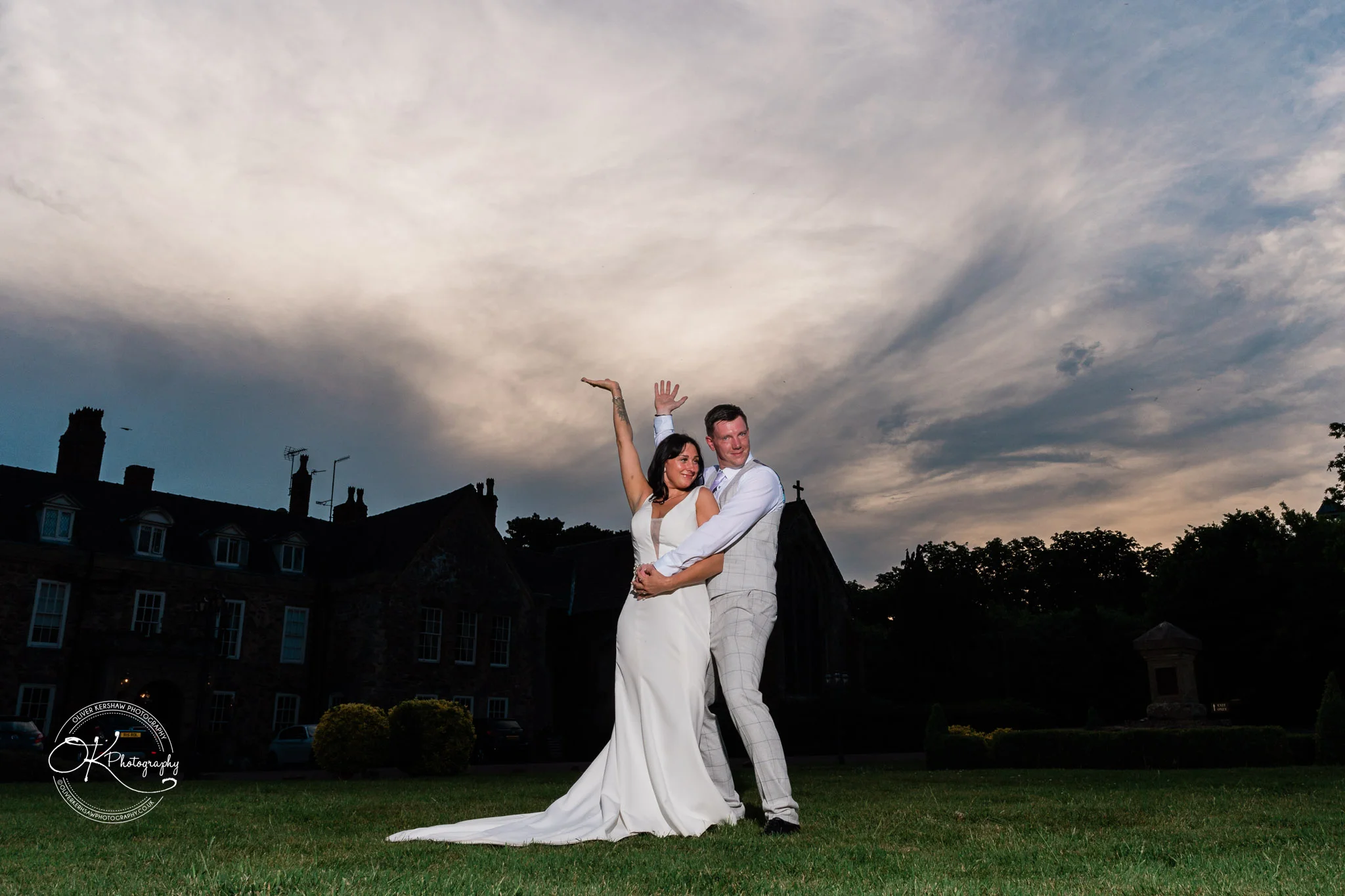 Bride and groom posing on a lawn with a dramatic evening sky background and a building in the distance.