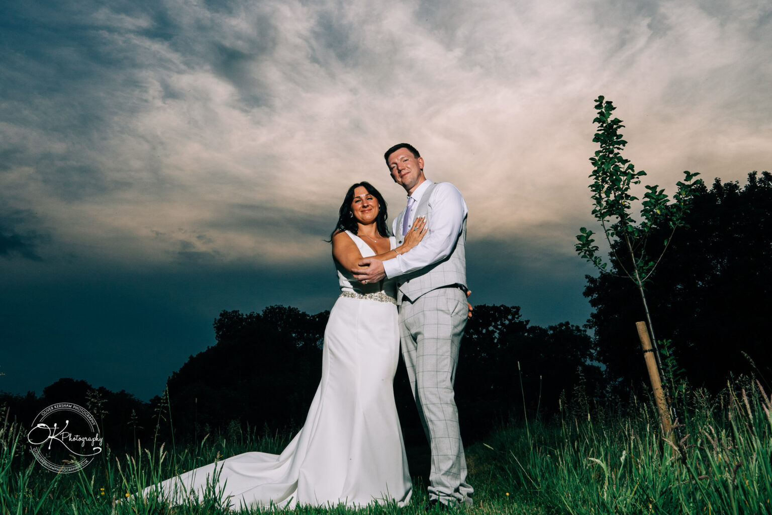 Rothley Court Hotel wedding photography A newlywed couple embracing in an open field with a dramatic sky in the background. The bride wears a white gown, and the groom wears a grey suit.
