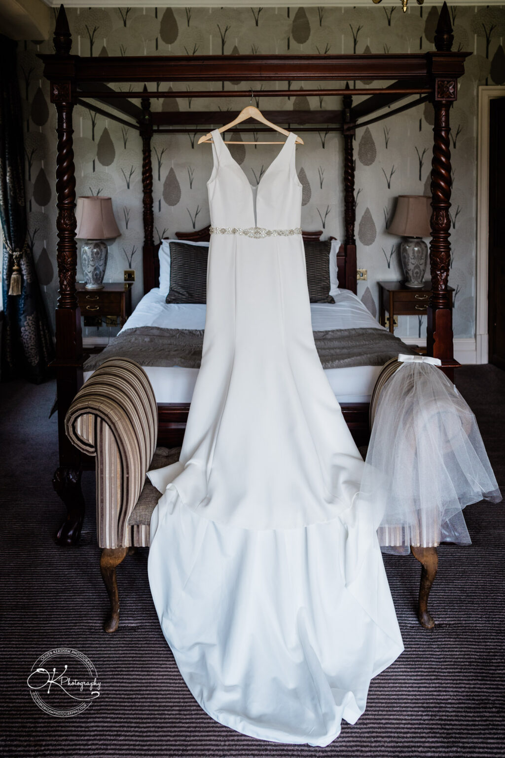 Rothley Court Hotel wedding photography White wedding dress with beaded belt hanging on a hanger from a four-poster bed, with a veil draped over a nearby striped chair.