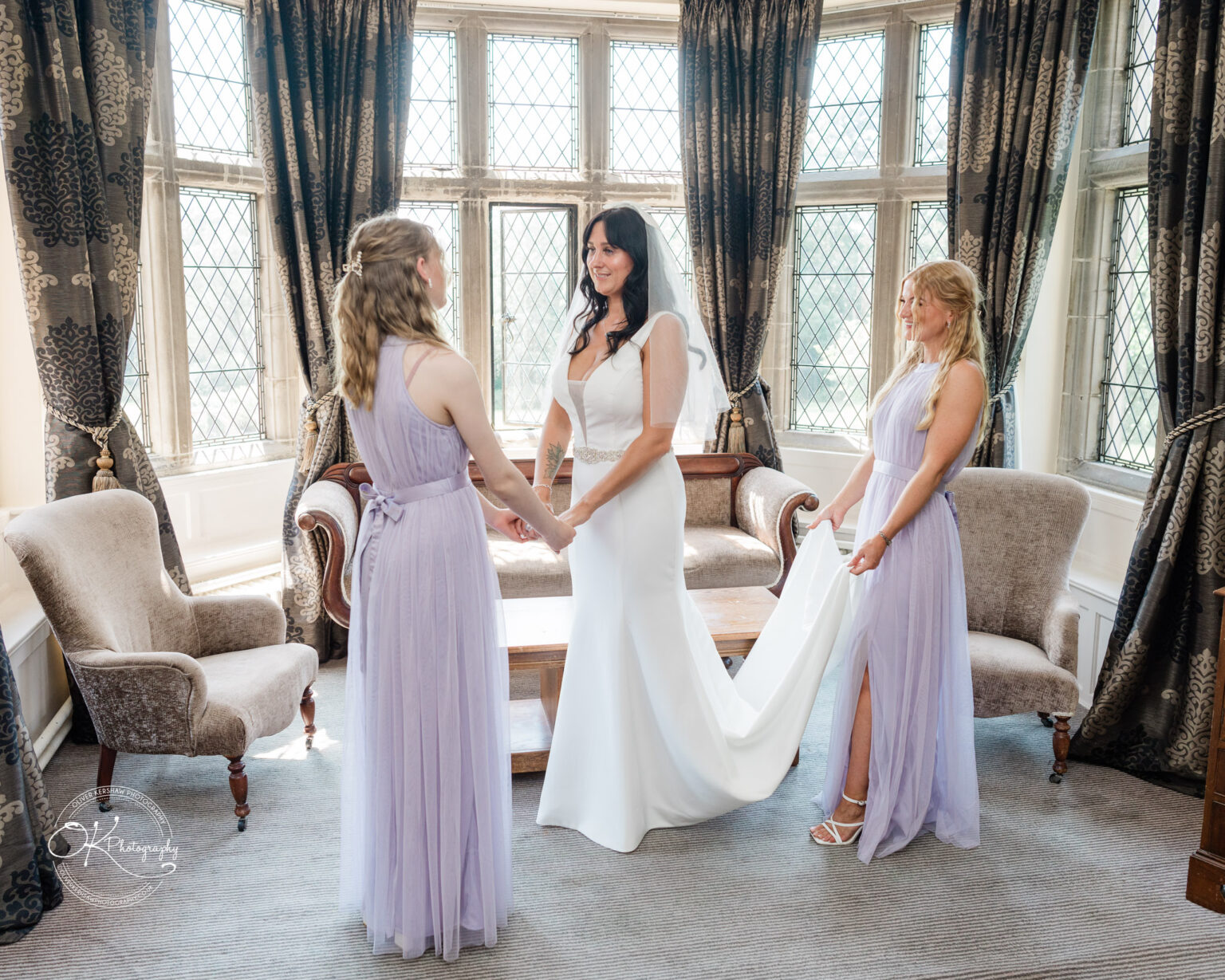 Rothley Court Hotel wedding photography A bride in a white dress stands between two bridesmaids wearing lavender dresses, in a room with large leaded windows and ornate curtains.