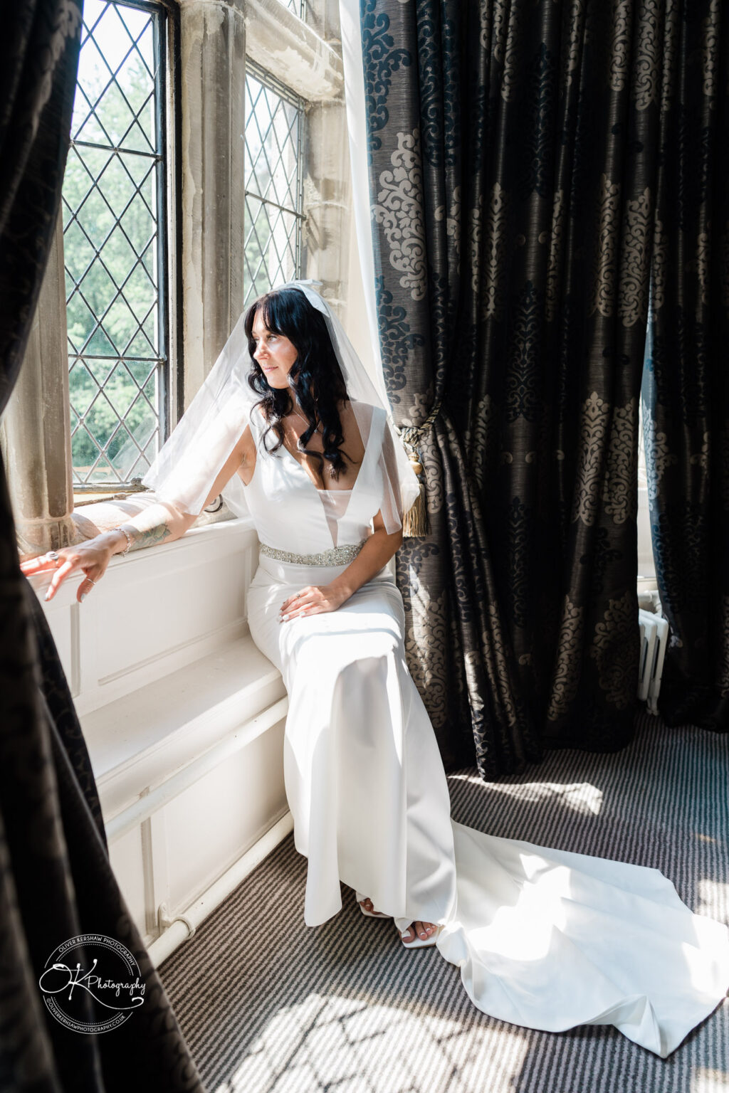 Rothley Court Hotel wedding photography Bride in a white dress and veil sitting on a window seat, looking out through diamond-paned windows with dark, patterned curtains.