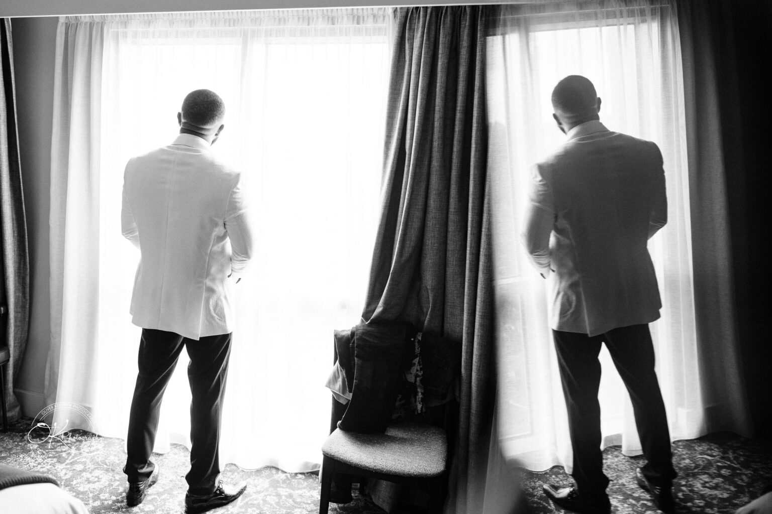 Symphony Rooms Wedding Photography A man in a white suit standing by a window with reflection on the glass, back facing the camera, in black and white.