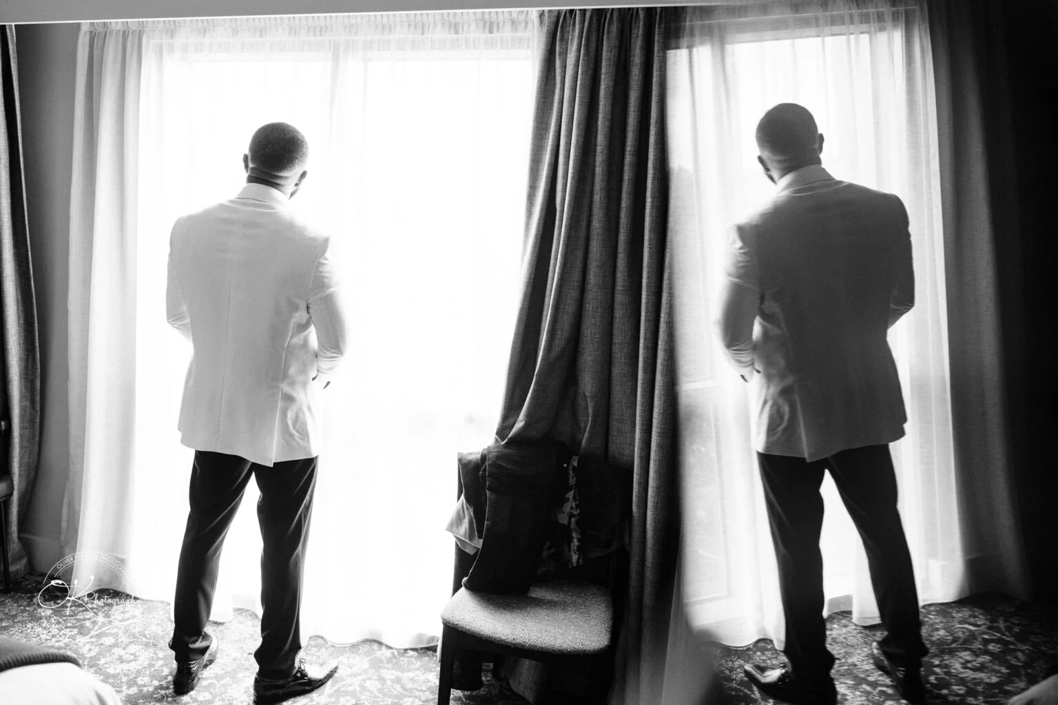A man in a white suit standing by a window with reflection on the glass, back facing the camera, in black and white.