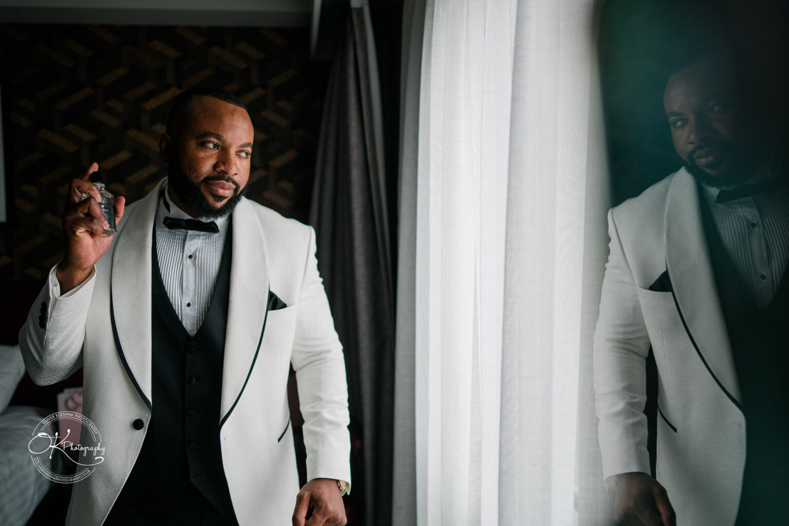 Symphony Rooms Wedding Photography Man in a white tuxedo spraying cologne and looking out of a window.