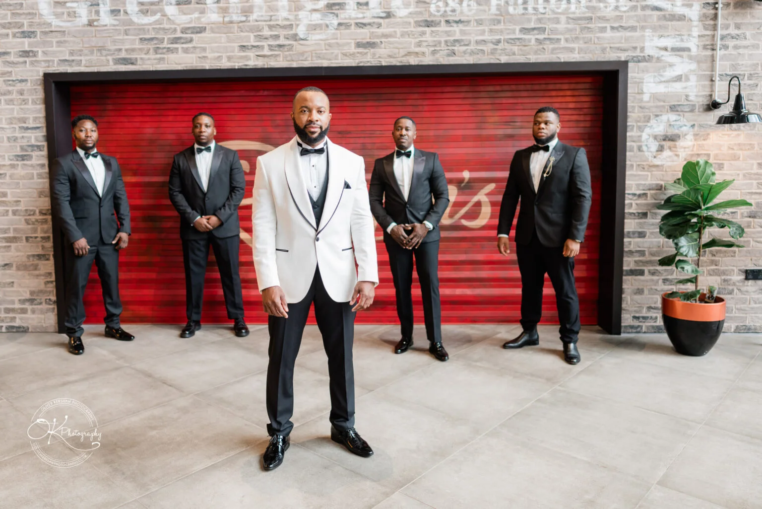 A group of five men dressed in formal suits, with one man in a white jacket standing in the foreground and the other four in black jackets standing behind him against a brick wall background.