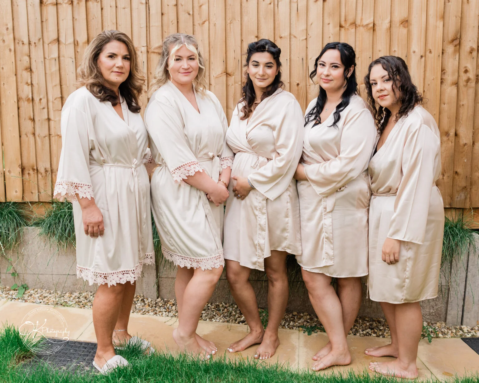 Five women in silk robes standing outdoors in front of a wooden fence.