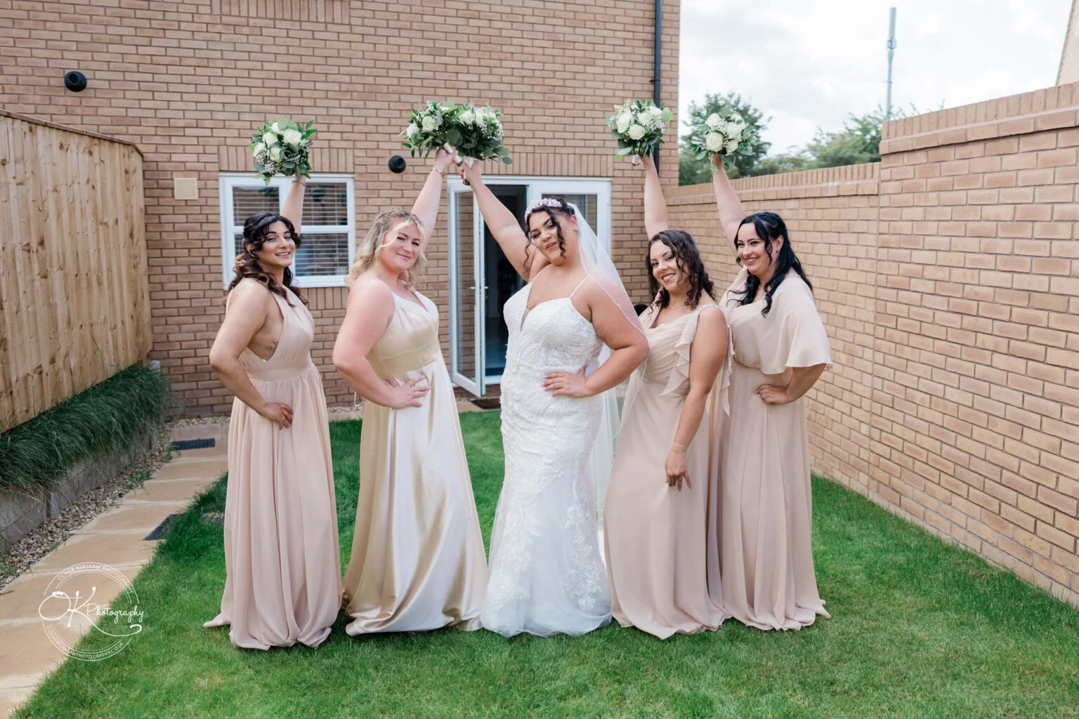 Bride in a white gown and veil with four bridesmaids in beige dresses, all holding bouquets aloft, standing in a garden with brick walls and a wooden fence.