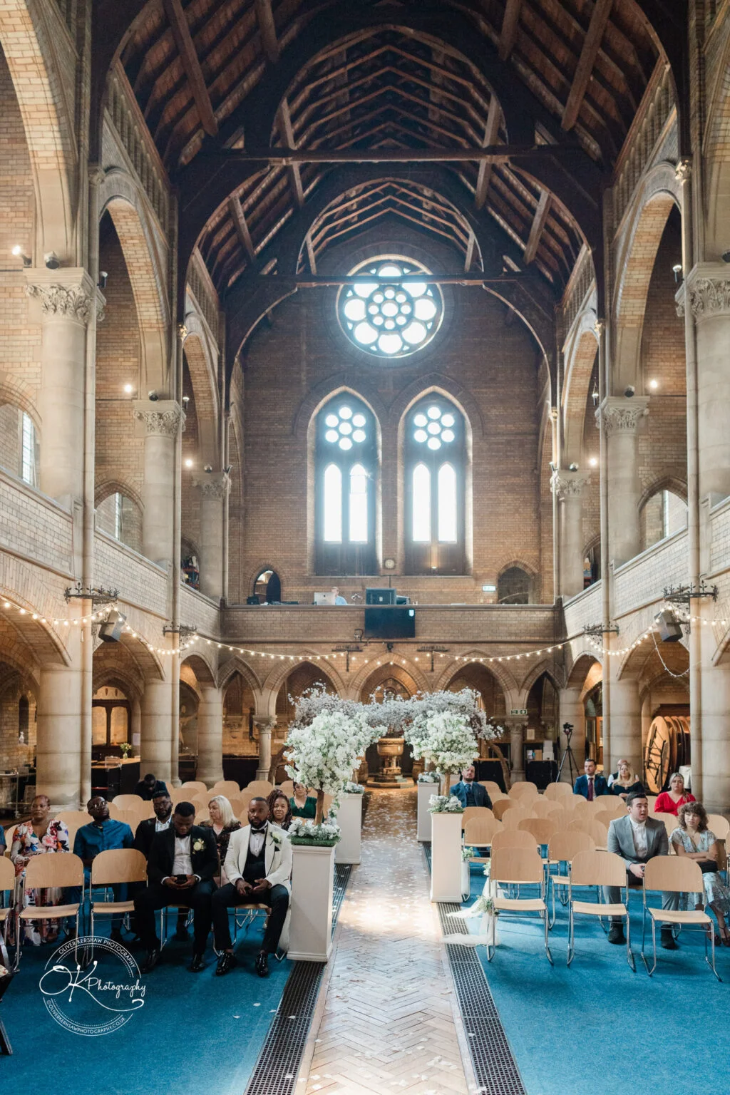 Interior of a grand church with high vaulted ceilings, large arched windows, and a central aisle decorated with white floral arrangements, filled with seated guests attending a ceremony.