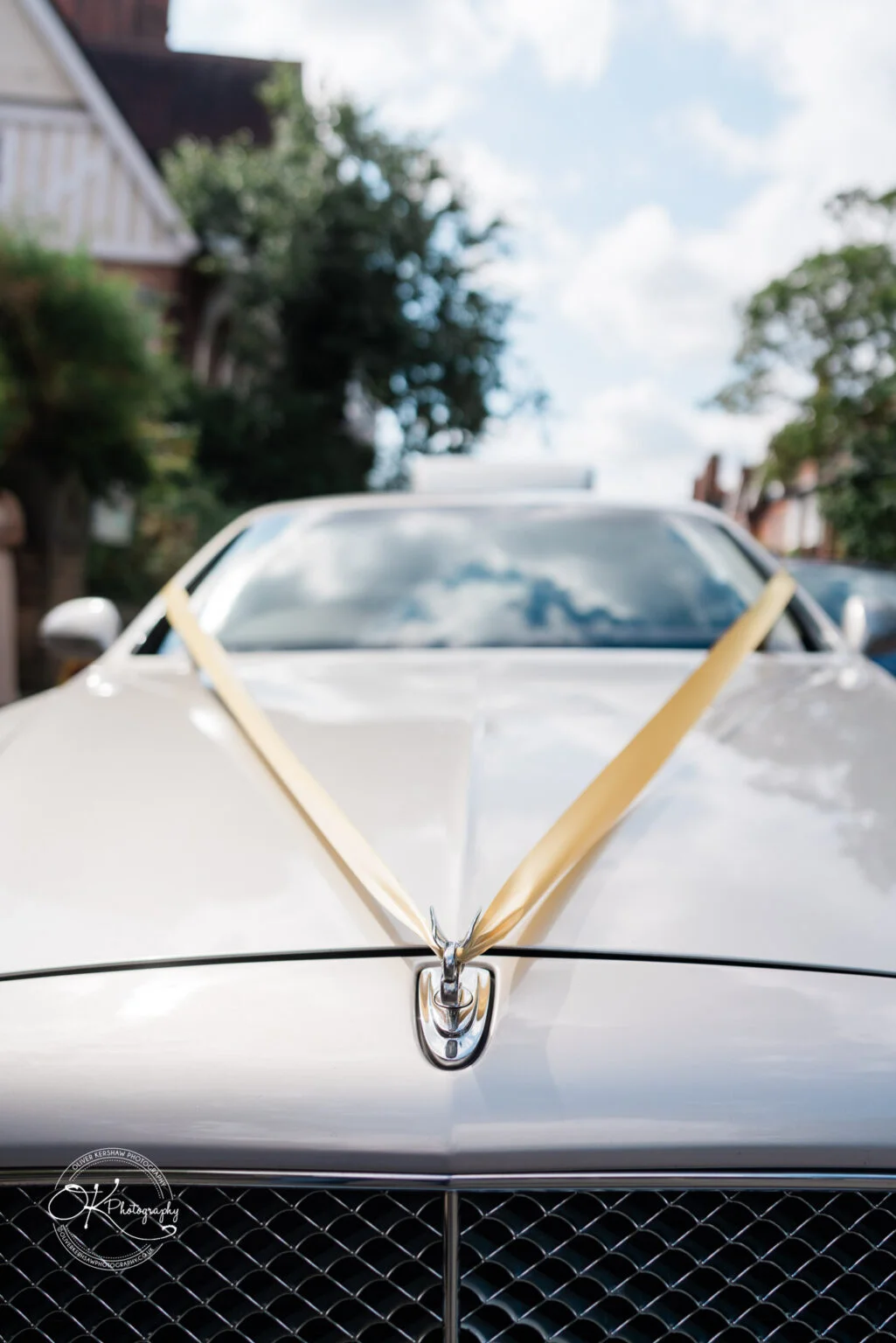 Front view of a silver luxury car with a golden ribbon tied across the hood, set in an outdoor residential area.