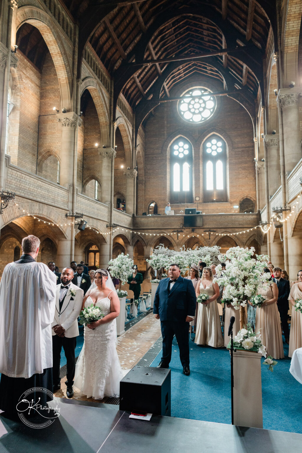 Symphony Rooms Wedding Photography A bride and groom stand at the altar during their wedding ceremony inside a large, ornate church with vaulted ceilings and stained glass windows. The bridal party and guests are seated on either side, surrounded by white floral arrangements.