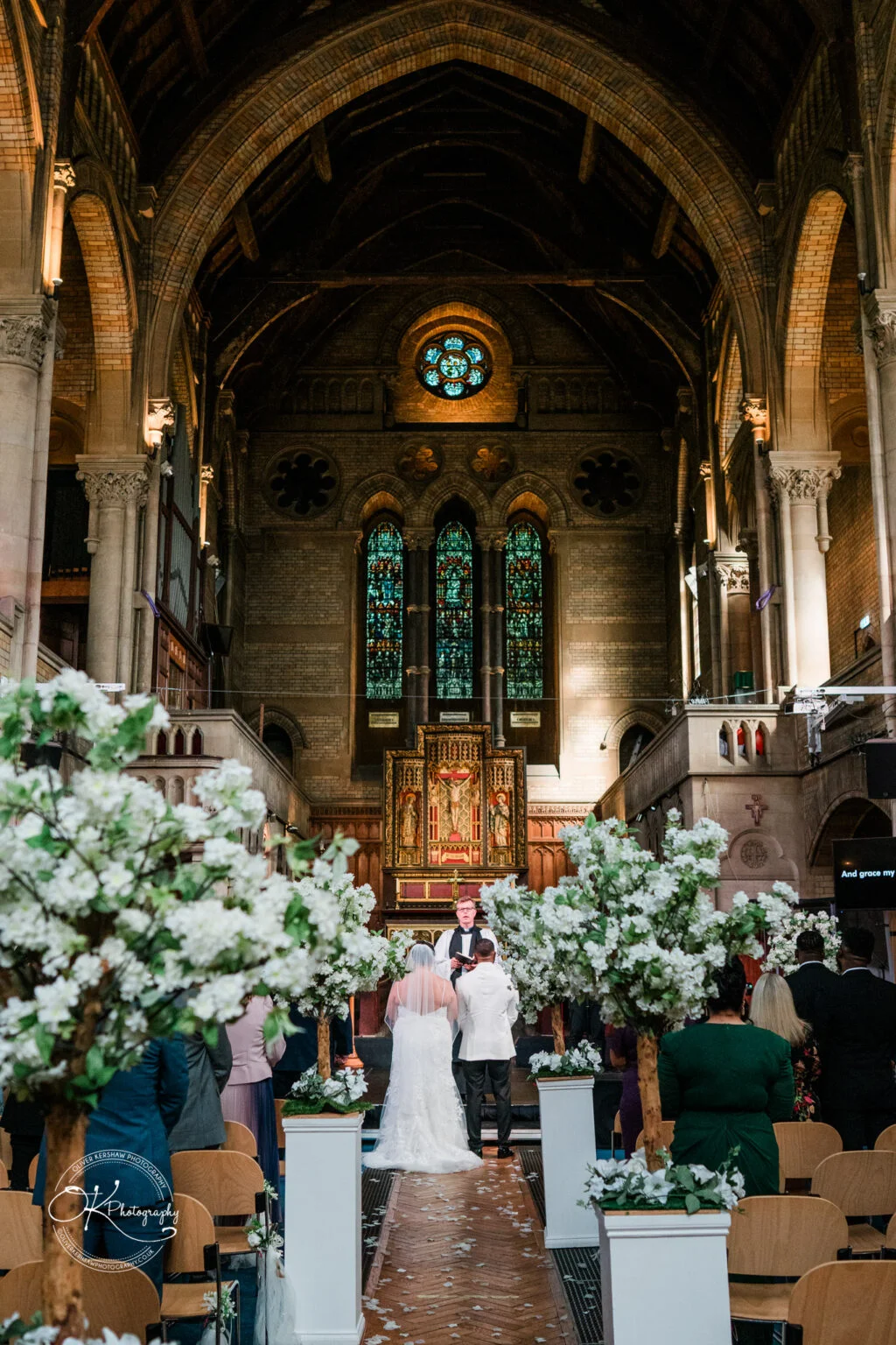 Bride and groom standing at the altar in a grand church with high arched ceilings, surrounded by wedding guests and white floral decorations.