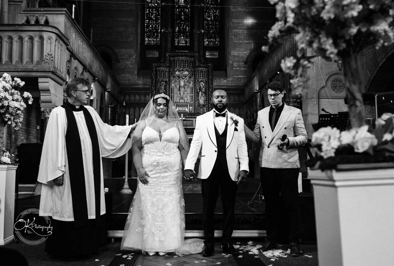 A bride and groom holding hands during a wedding ceremony in a church, flanked by two officiants.