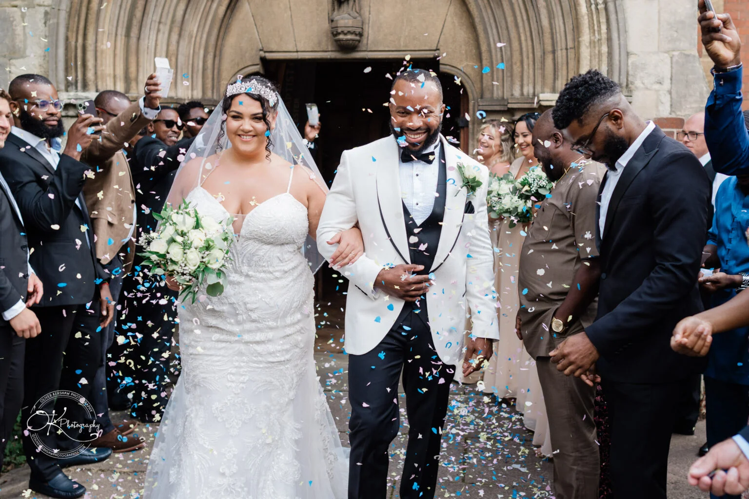 Bride and groom smiling and walking arm-in-arm among wedding guests throwing confetti in front of a church.