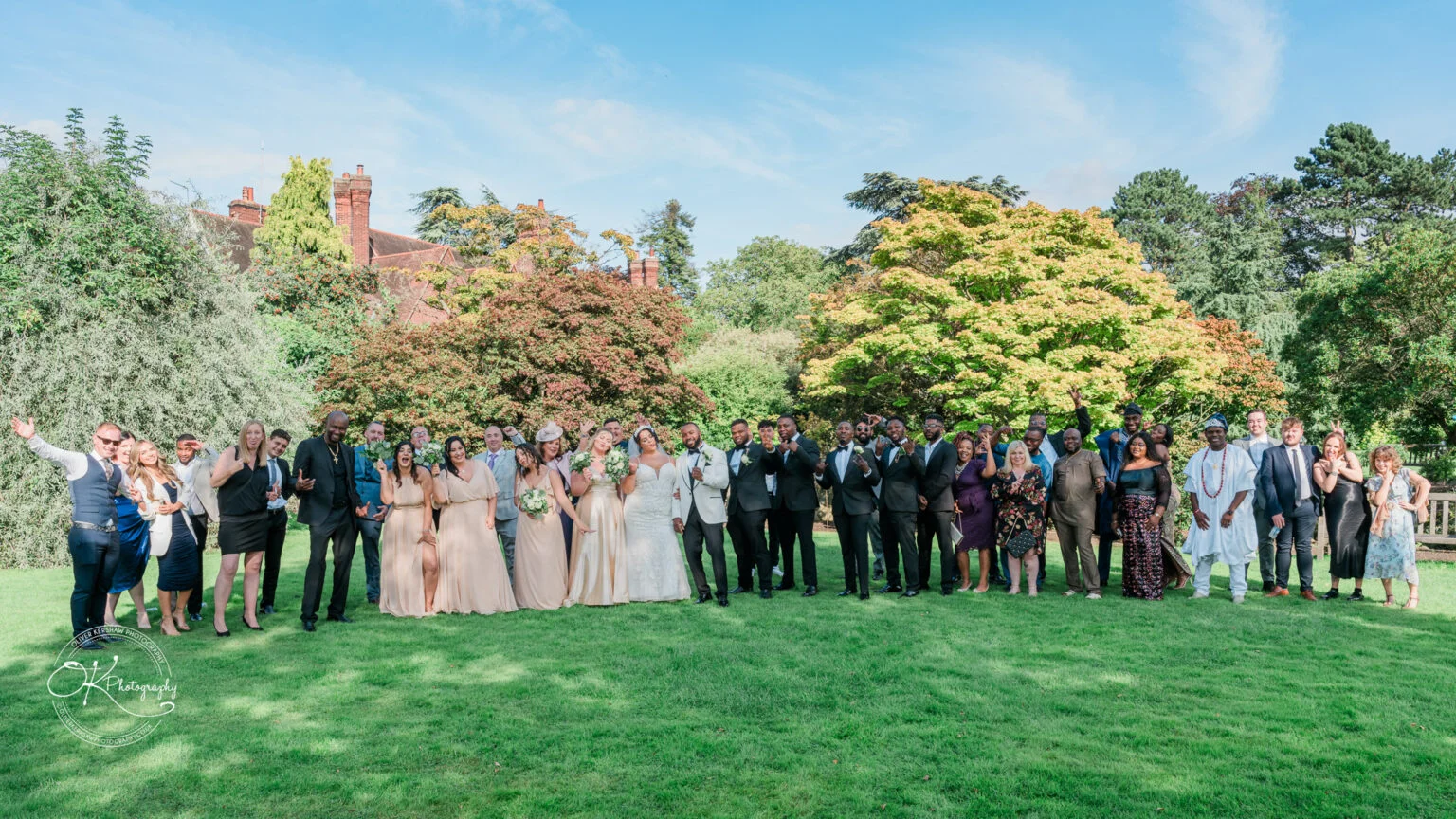 A large wedding party posing for a group photo outdoors in front of trees and a building, with the bride and groom in the centre.