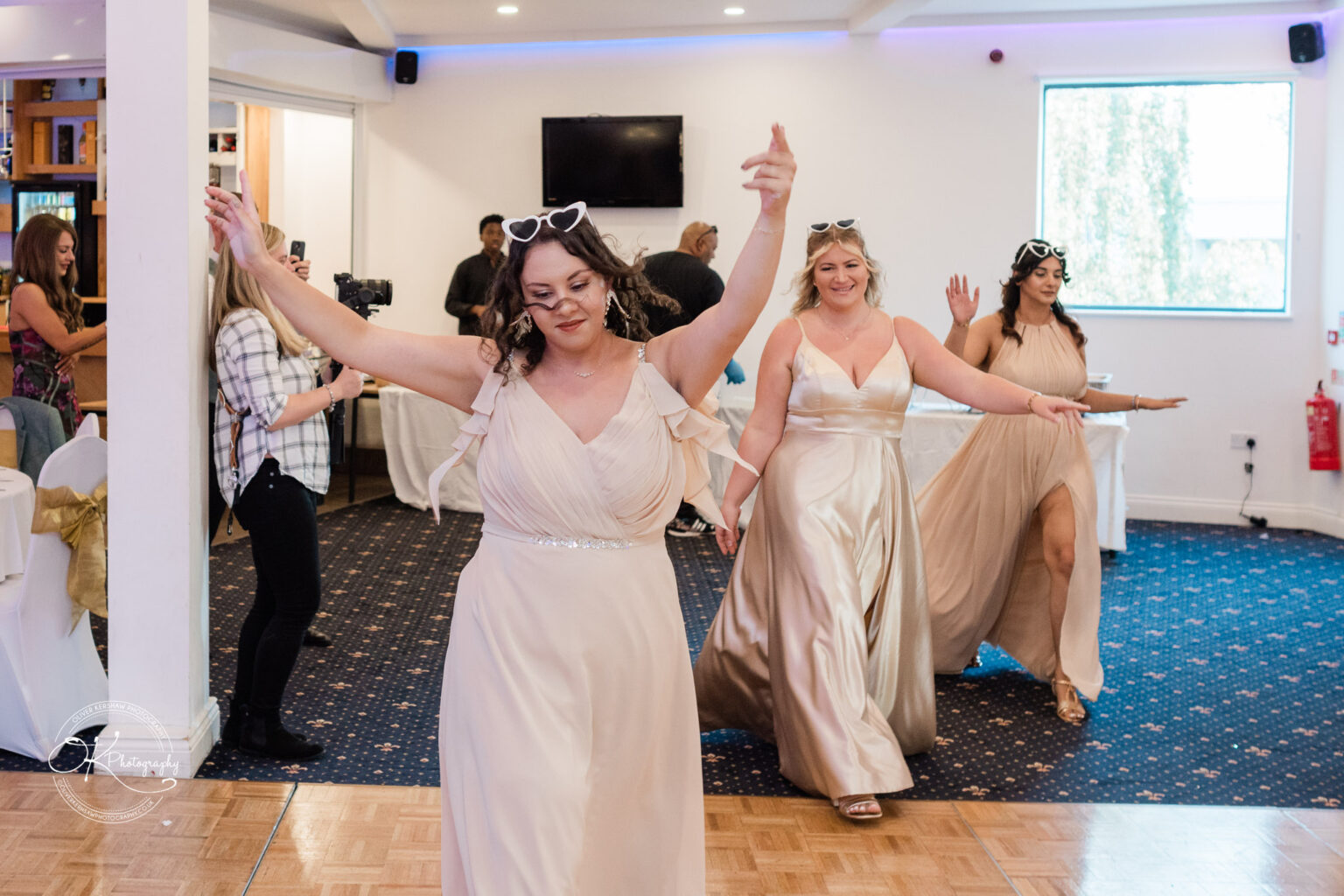 Symphony Rooms Wedding Photography Three women in flowing beige dresses and heart-shaped sunglasses dance in a well-lit room with other guests in the background.