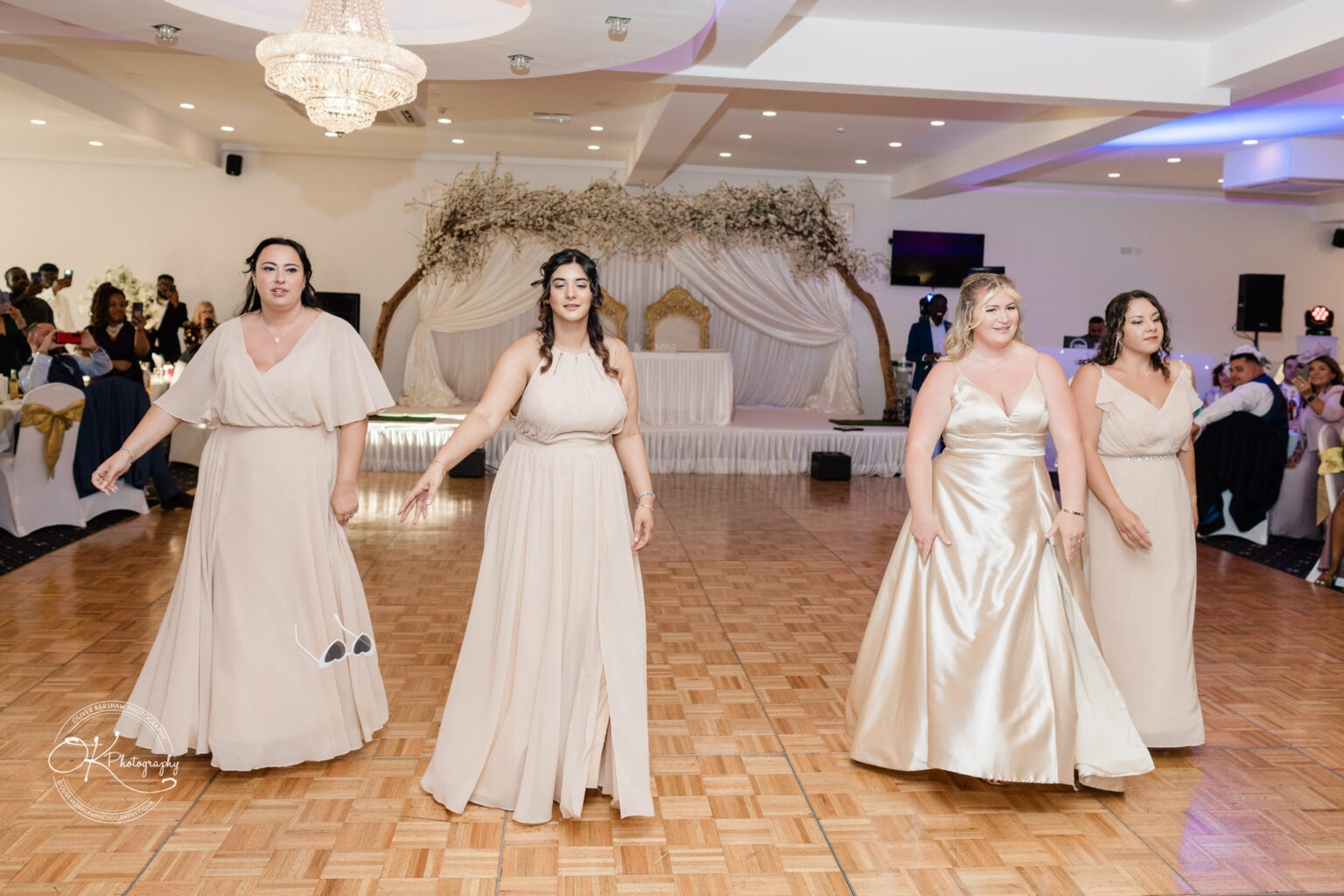 Four bridesmaids in beige dresses walking on a wooden dance floor at a wedding reception.