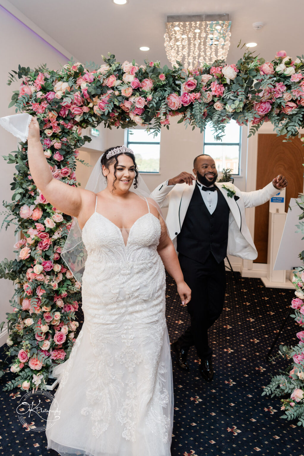 Symphony Rooms Wedding Photography A bride and groom walk under a floral arch adorned with pink and white roses and greenery, both looking joyful, with the bride holding up her bouquet.