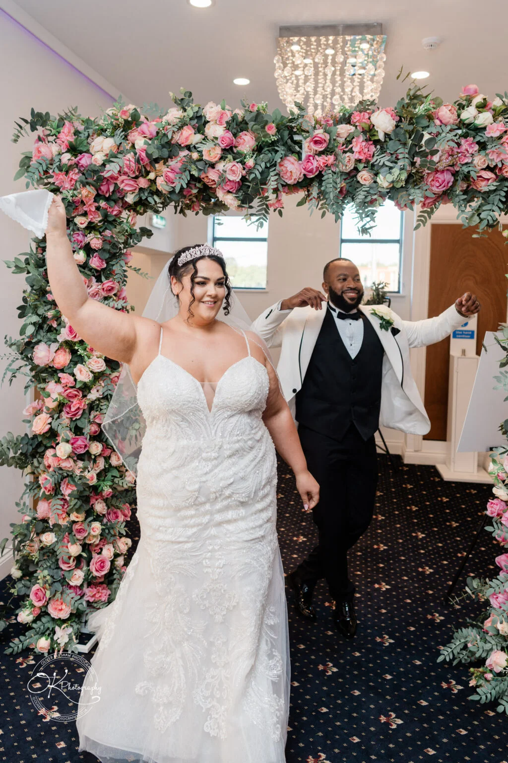 A bride and groom walk under a floral arch adorned with pink and white roses and greenery, both looking joyful, with the bride holding up her bouquet.
